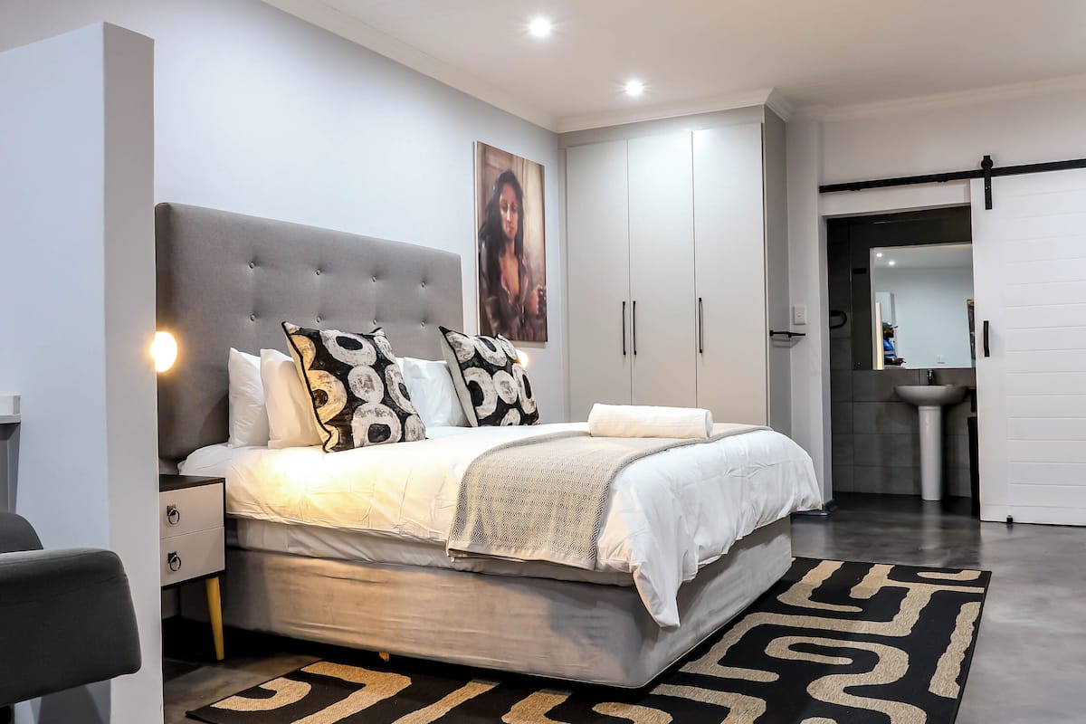 Une chambre moderne avec une tête de lit capitonnée grise, un grand lit agrémenté d’oreillers à motifs, une table de chevet et une armoire à portes coulissantes.
