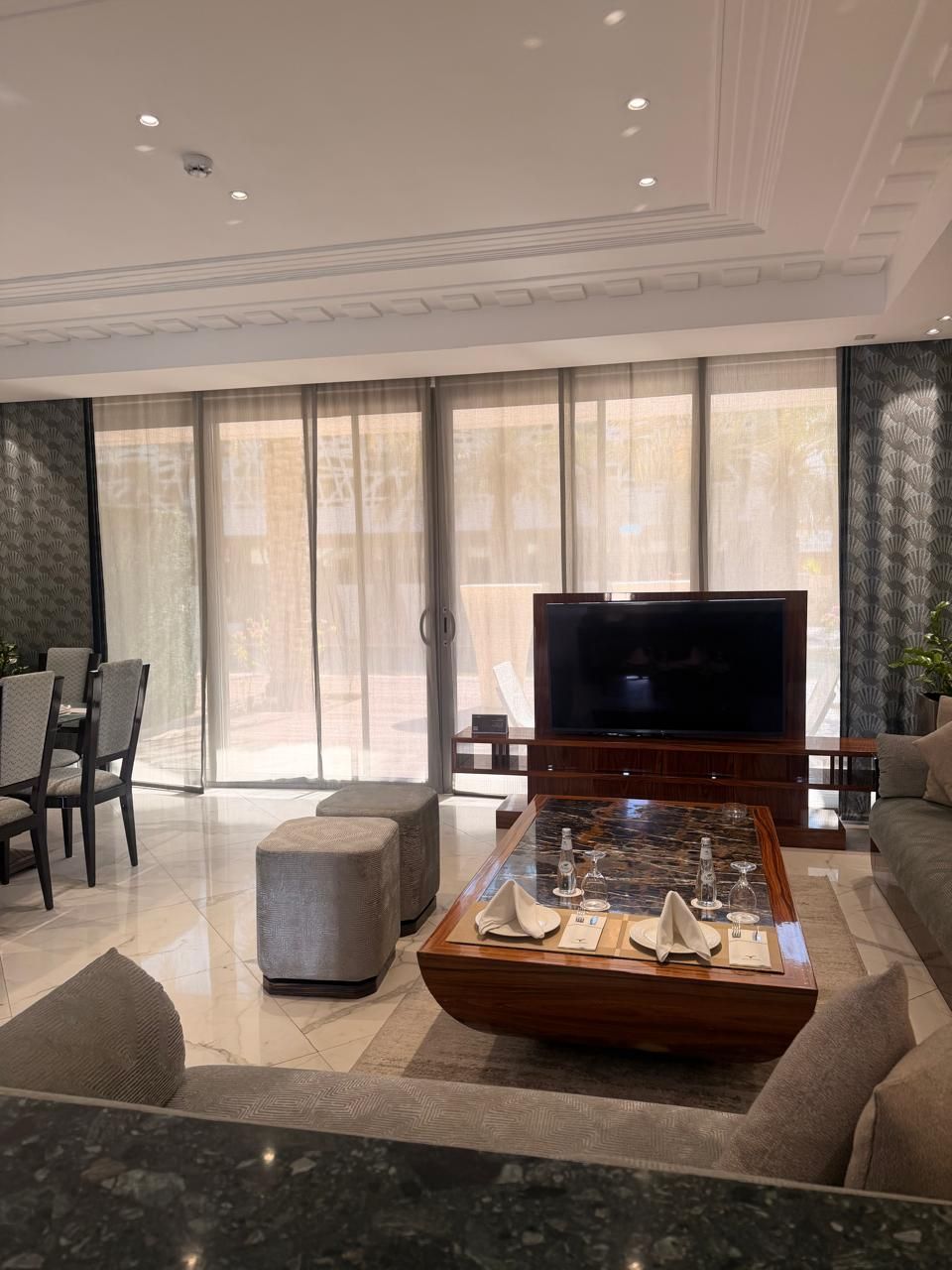 Una sala de estar moderna con un televisor de pantalla plana, una mesa de centro de vidrio con servilletas, y un área de estar con un sofá y sillones.