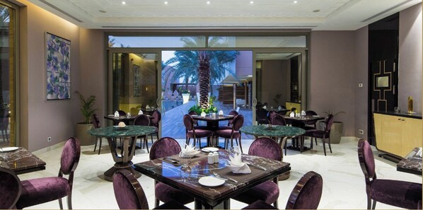 A dining area with multiple tables and chairs, a large window with a view of palm trees, and a modern interior design. - Vivienda Hotel Villas Al Hada (Riyadh)