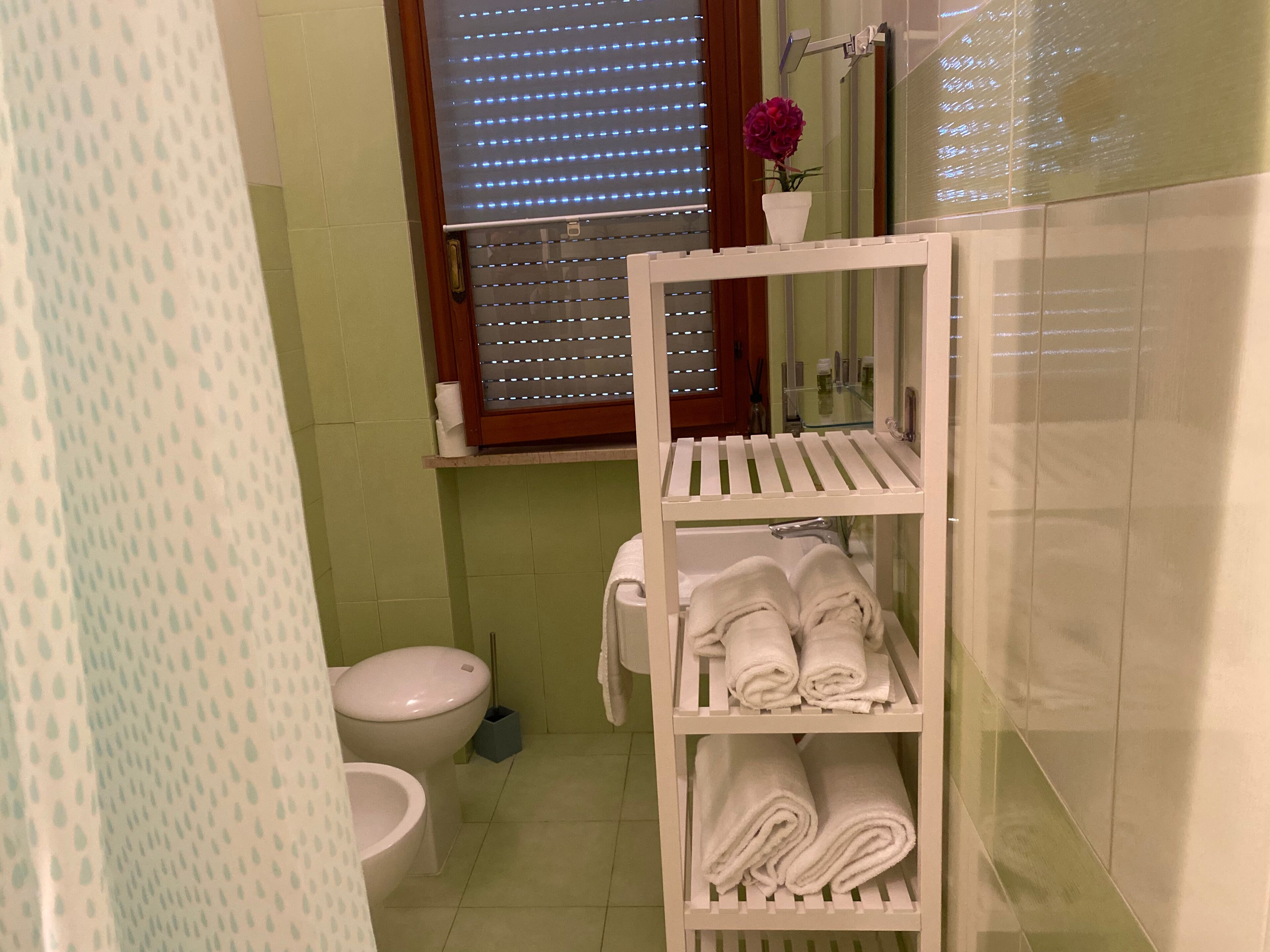 Un baño con inodoro, lavabo, estante con toallas y ventana con persianas.