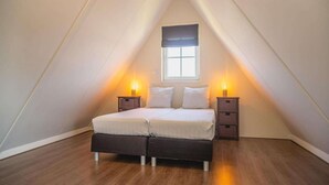Un dormitorio con techo inclinado, una cama con dos almohadas, dos mesitas de noche con lámparas y un ventanal con diseño de cruz.