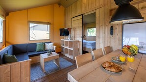 Interior acogedor de una cabaña con una mesa de madera dispuesta para el desayuno, un sofá con cojines, un televisor en un estante y un gran ventanal con vistas al exterior.