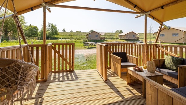 Una terraza de madera con una silla hamaca, un banco acolchado y una mesa con una cesta y vasos.