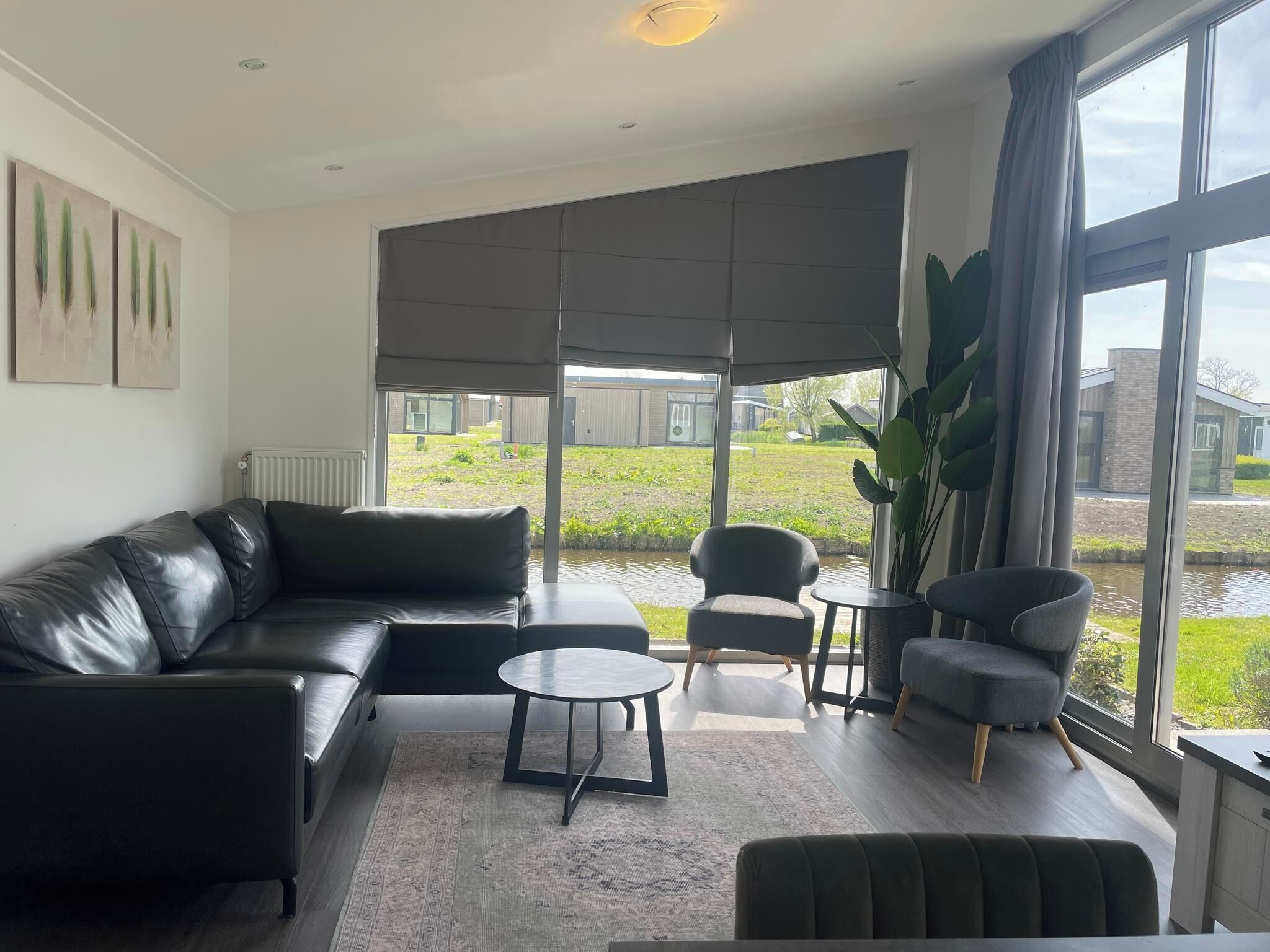 Un salon moderne avec un canapé en cuir, une table basse ronde et deux fauteuils. Une grande fenêtre offre une vue sur une cour intérieure agrémentée d’un élément aquatique et sur des bâtiments en arrière-plan.