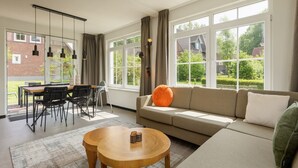 Een moderne woonkamer met een bank, een eettafel en stoelen. Grote ramen bieden uitzicht op het exterieur.