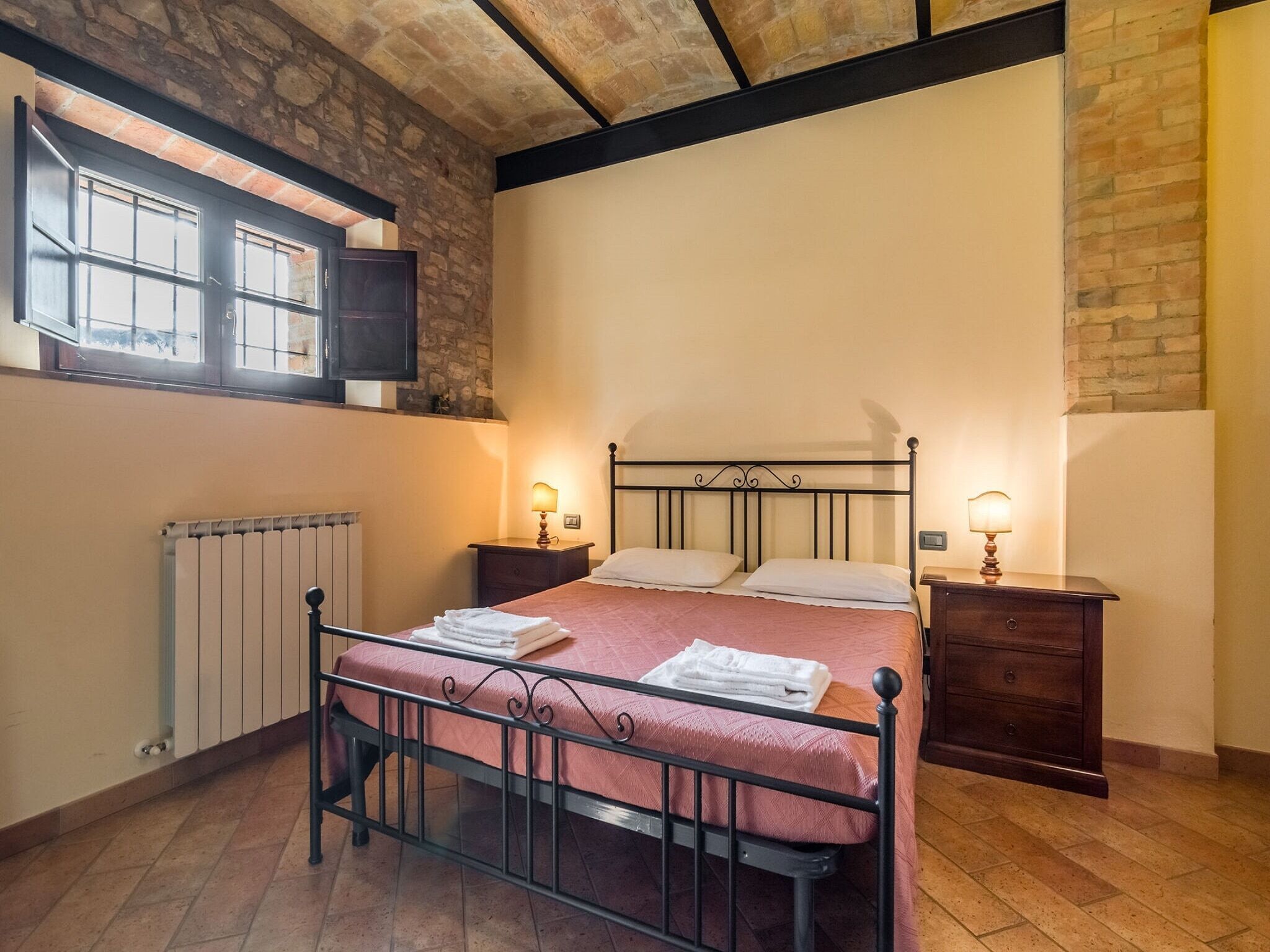 A bedroom with a bed, two bedside tables, a window, a radiator, and a brick wall.