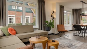 A modern living room with a sofa, two wooden side tables, a potted plant, a dining area with chairs, and a view of a brick building through large windows. - Home w/ Sauna & Garden (Bad Bentheim)