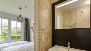 Hotelkamer met een bed, een raam met gordijnen, een hanglamp, een badkamer met een wastafel en spiegel, en een betegelde wand.