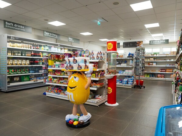 A shopping area with a large mascot figure, shelves stocked with various products, and signs indicating different sections like 'Soft drinks' and 'Eggs & Dairy.'