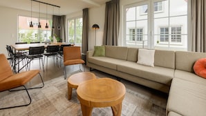 En moderne stue med en sofa, to træsideborde, et spisebord med stole og et stort vindue med gardiner.