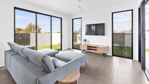 Un salon moderne avec un canapé gris, un téléviseur fixé au mur et de grandes portes-fenêtres coulissantes donnant sur un jardin.