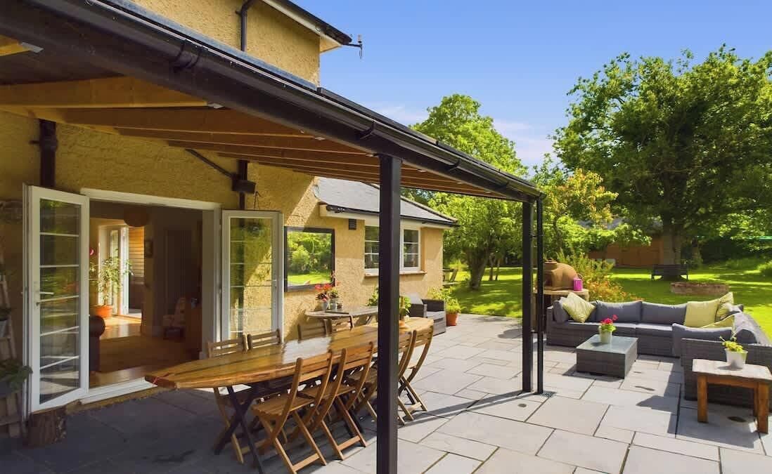 Une terrasse couverte avec une table et des chaises en bois, un canapé et une petite table sur laquelle est posé un vase.