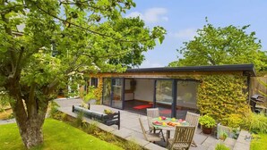 A modern house with a glass-enclosed patio, a dining table with chairs, and a garden with a tree and plants.