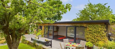 A modern house with a glass-enclosed patio, a dining table with chairs, and a garden with a tree and plants.