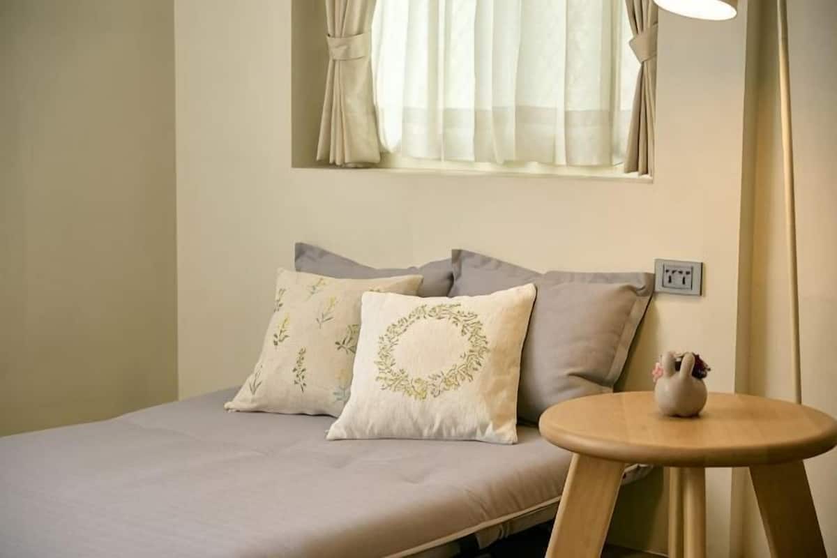 A bed with pillows, a small table, a window with curtains, and a wall socket.