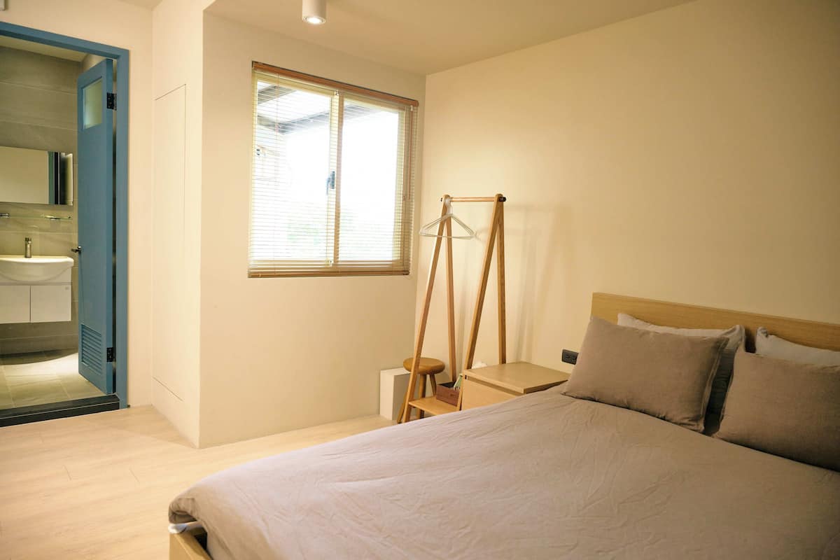 A bedroom with a bed, a wooden headboard, a bedside table, a hanging clothes rack, and a window with blinds.