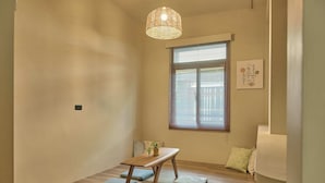 Et værelse med trægulv, et lille bord med bænk, en hængende lampe, et vindue med persienner og et billede hængt på væggen.