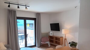 Ein modernes Wohnzimmer mit einer Couch, einem kleinen Tisch, einem Holzesstisch, einem an der Wand montierten Fernseher und einer Topfpflanze.