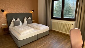 Ein ordentlich bezogenes Bett mit grauem Kopfteil und weißer Bettwäsche, ein Nachttisch aus Holz mit einer Lampe, ein Fenster mit Vorhängen und ein Heizkörper.