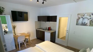 A compact hotel room with a bed, a desk, a kitchenette, and a door with a mirror.