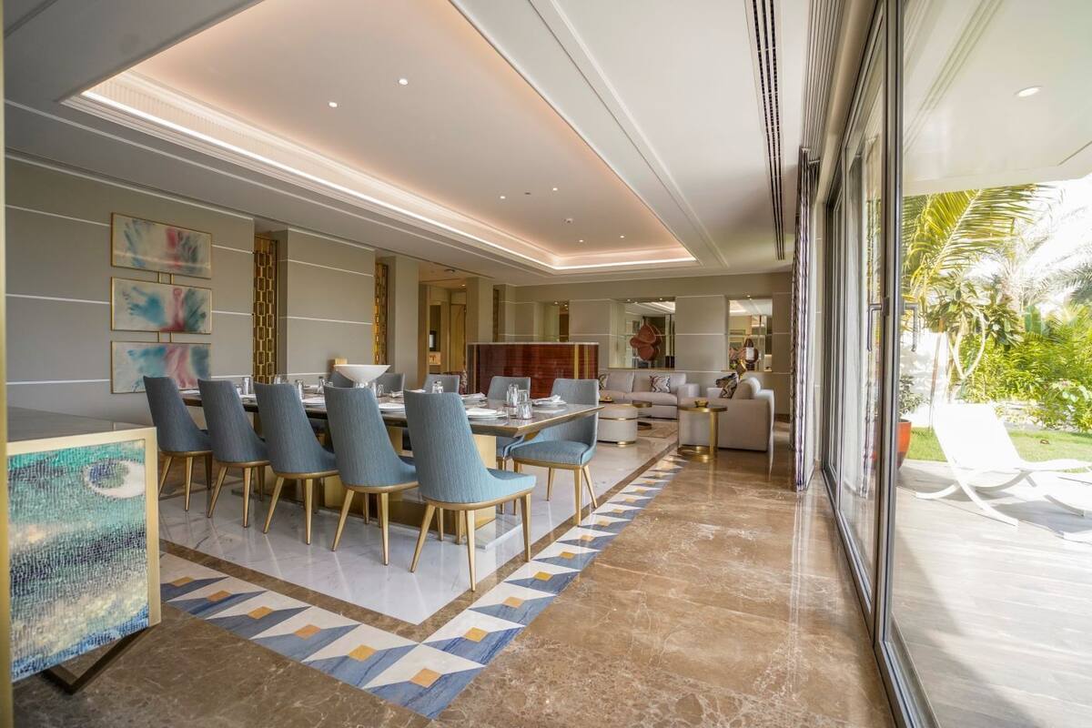 A modern dining area with a long table and chairs, a kitchen in the background, and a view of a garden through large windows.