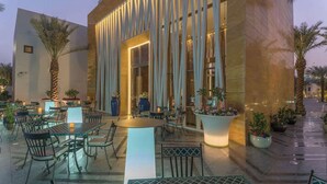 An outdoor dining area with tables and chairs, a large planter with flowers, and a building with vertical light fixtures. - Vivienda Hotel Villas Jeddah (Jeddah)