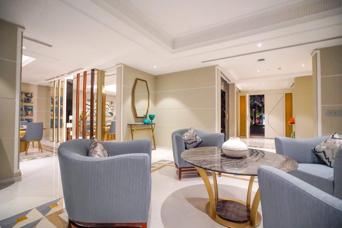 A modern hotel lobby with a round marble table, two armchairs, and a dining area with a glass partition.