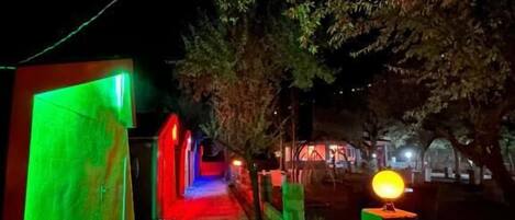 Un sendero estrecho con iluminación verde y roja, bordeado por muros y un árbol.