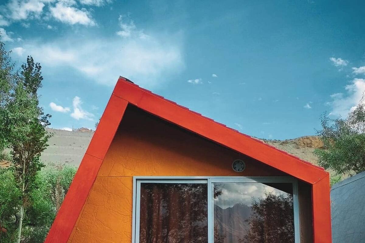 A small, triangular building with a red roof and orange walls, featuring two rocking chairs with striped cushions in front of a glass door.