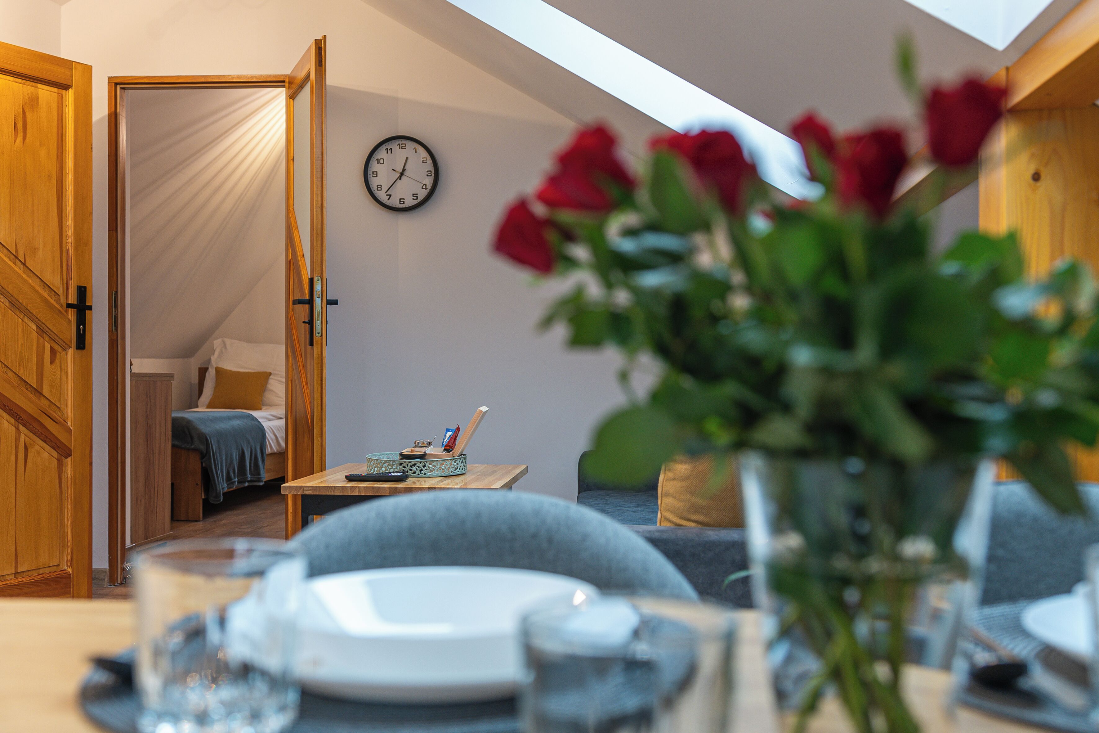 Une pièce confortable avec un lit, une horloge au mur et une table sur laquelle est posé un vase rempli de roses rouges.