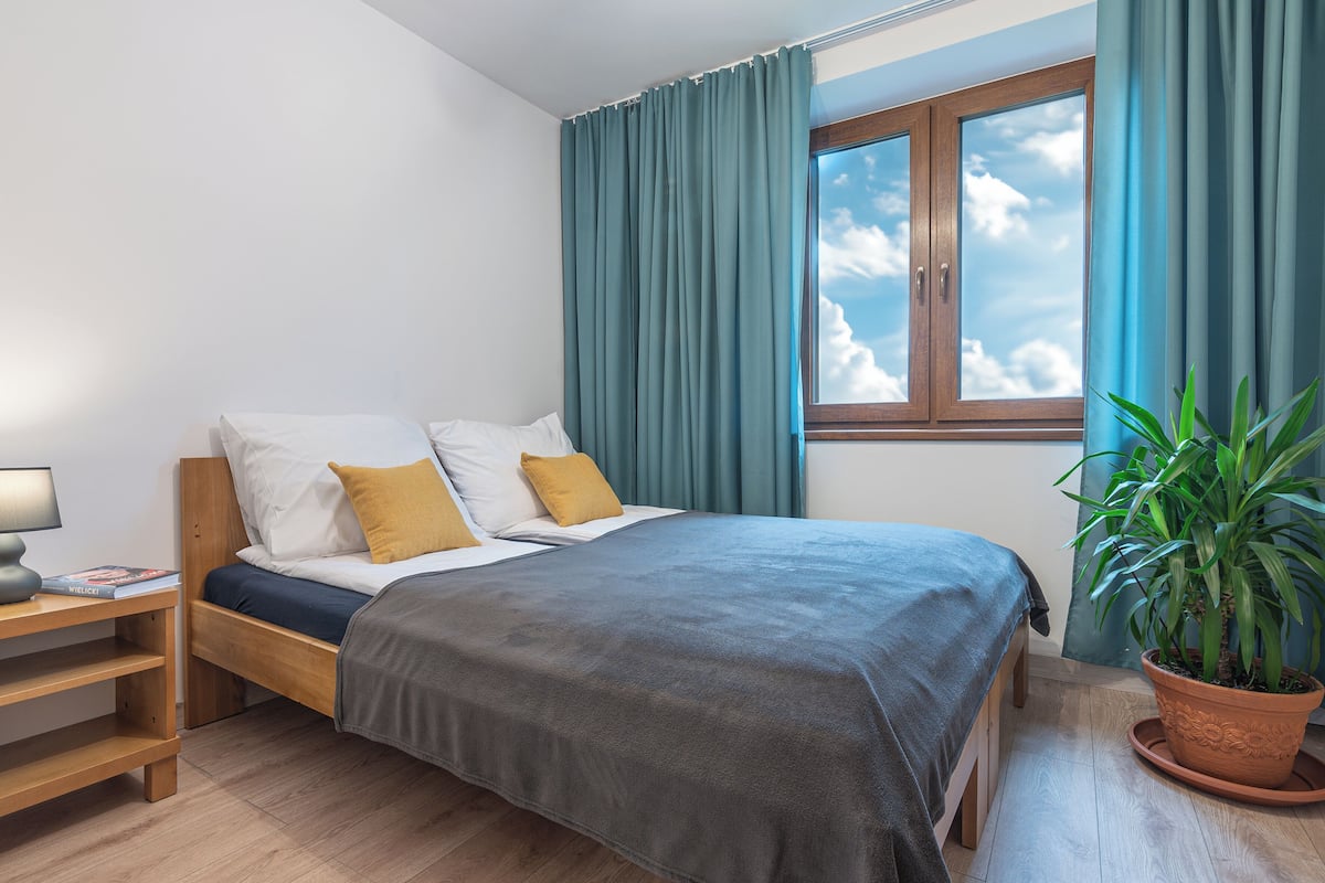 Ein Schlafzimmer mit Bett, Nachttisch, einem Fenster mit Vorhängen und einer Topfpflanze.