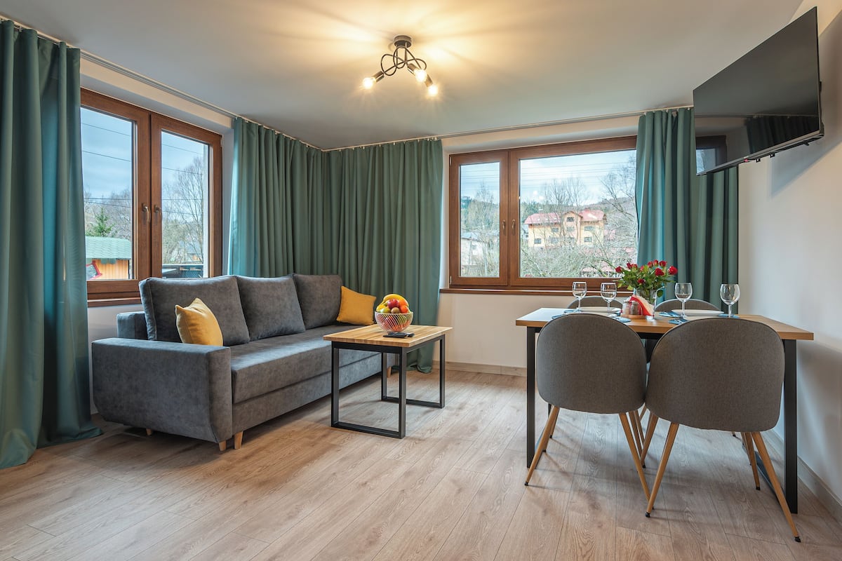 Ein modernes Wohnzimmer mit einer Couch, einem Esstisch mit Stühlen, einem an der Wand montierten Fernseher und großen Fenstern mit Vorhängen.