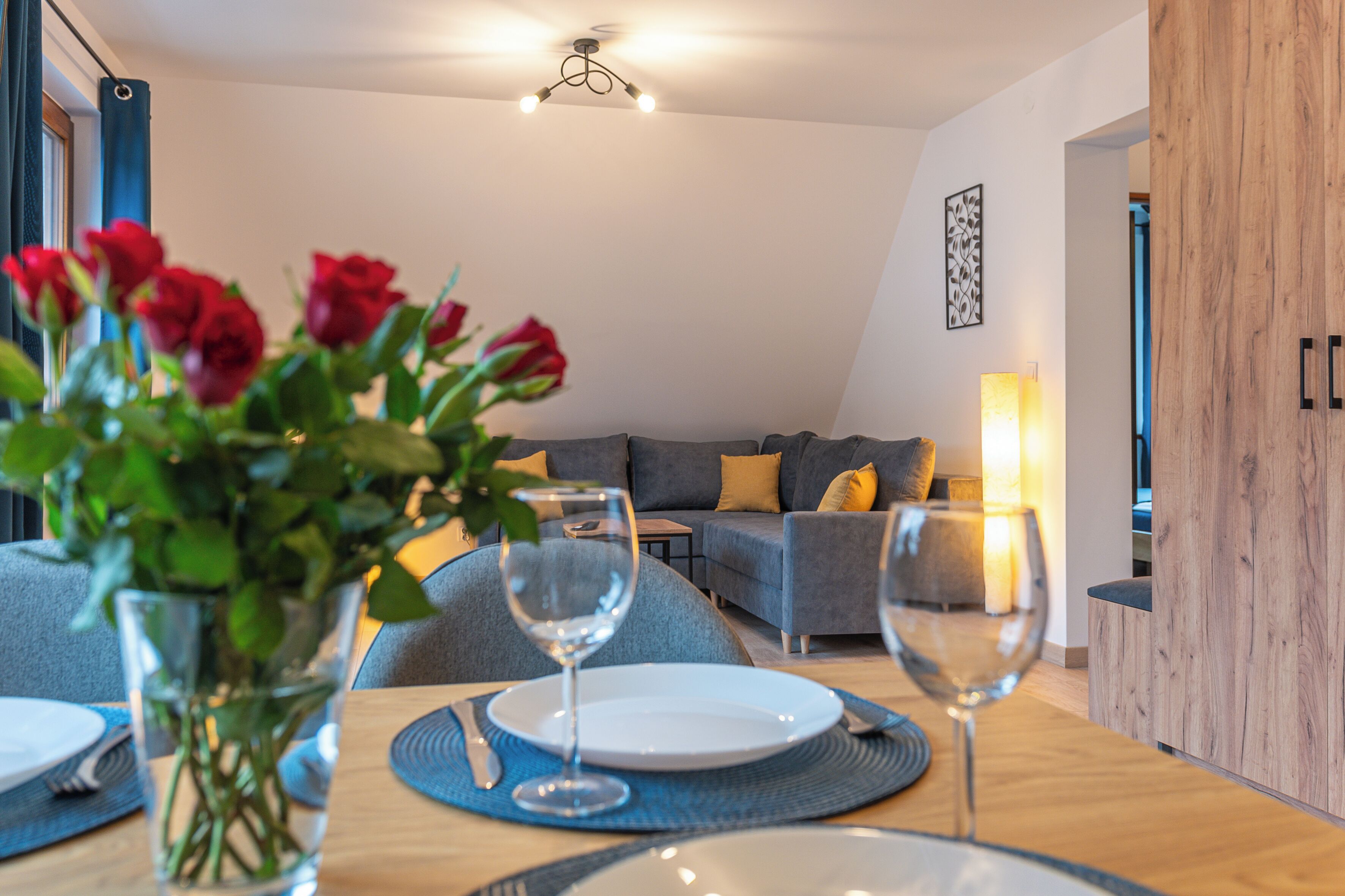 A dining area with a table set for two, a vase of red roses, and a modern living space in the background.