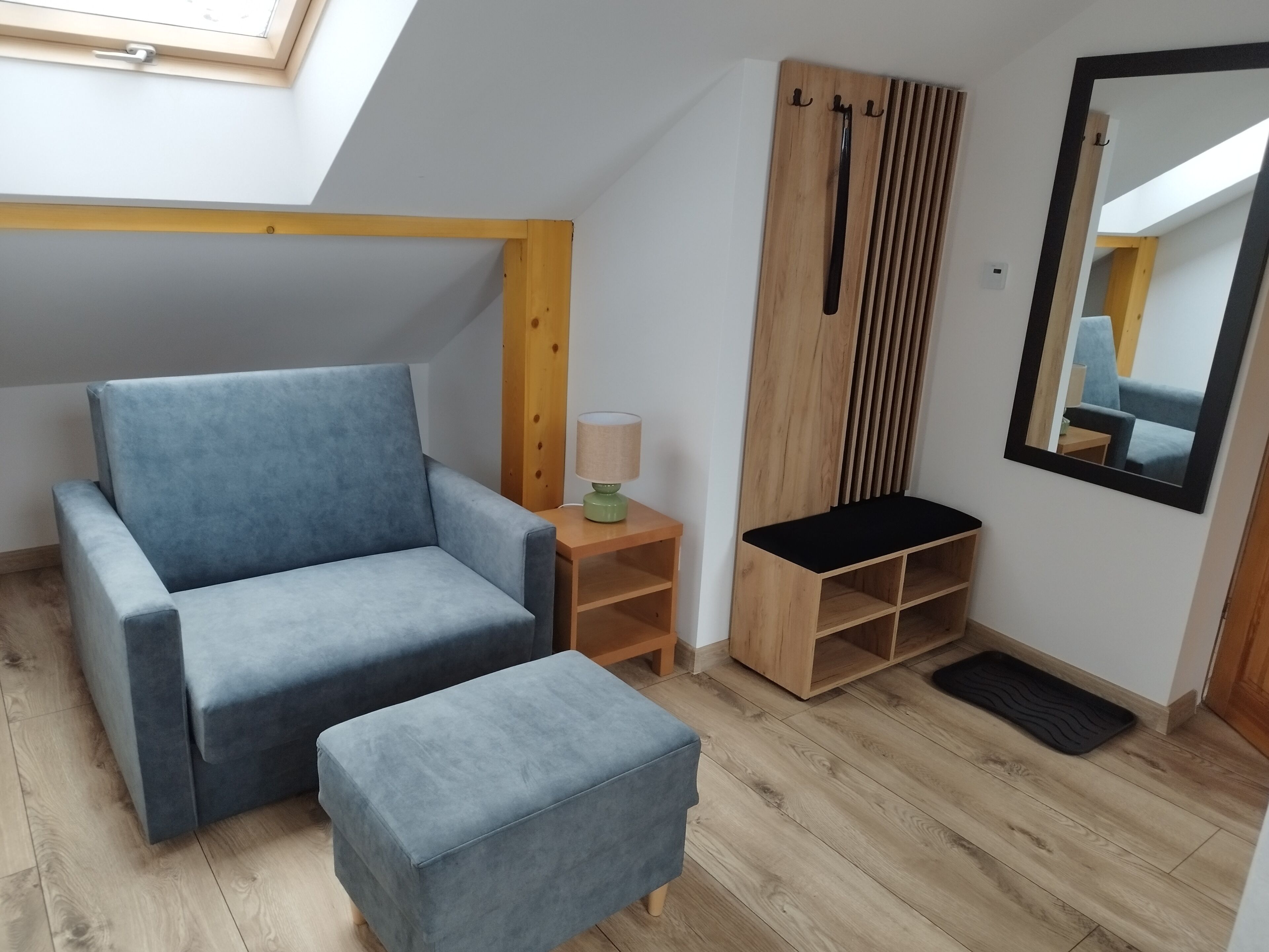 A room with a blue sofa, a matching ottoman, a wooden side table with a lamp, a black shelf, a wooden wardrobe, and a mirror on the wall.