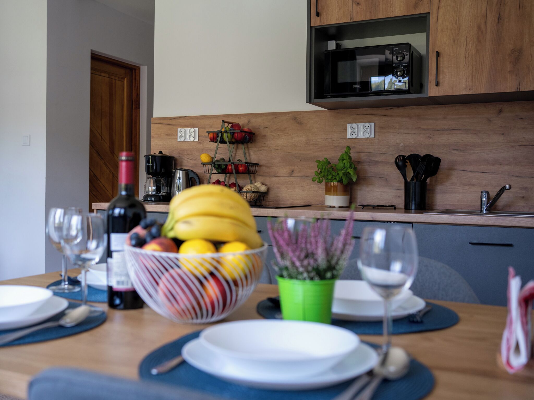 Una mesa de comedor con un frutero, copas de vino y platos. Un microondas está sobre la encimera, y hay armarios y una planta en la pared.