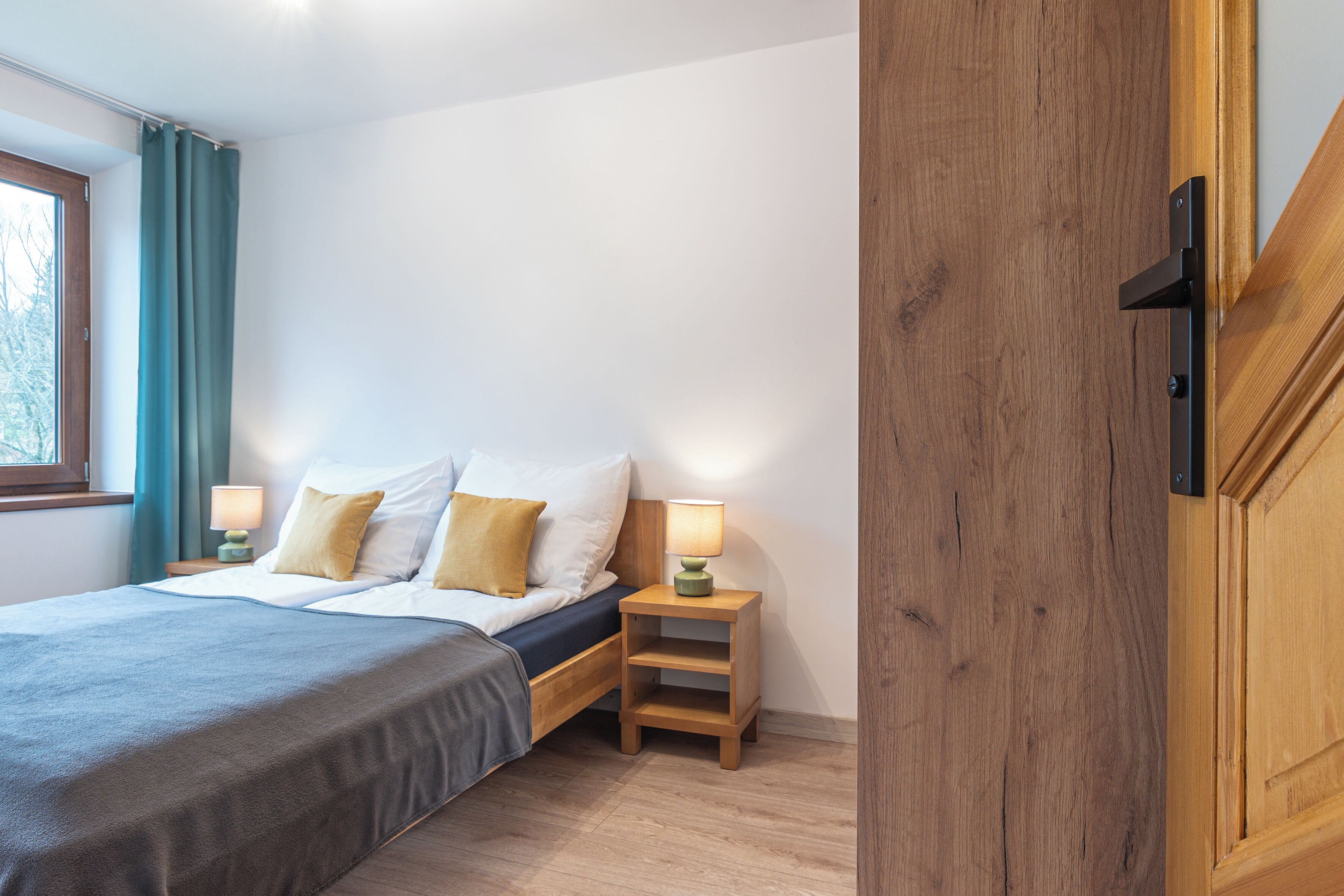 A bedroom with a bed, nightstands, a wooden door, and a window with curtains.