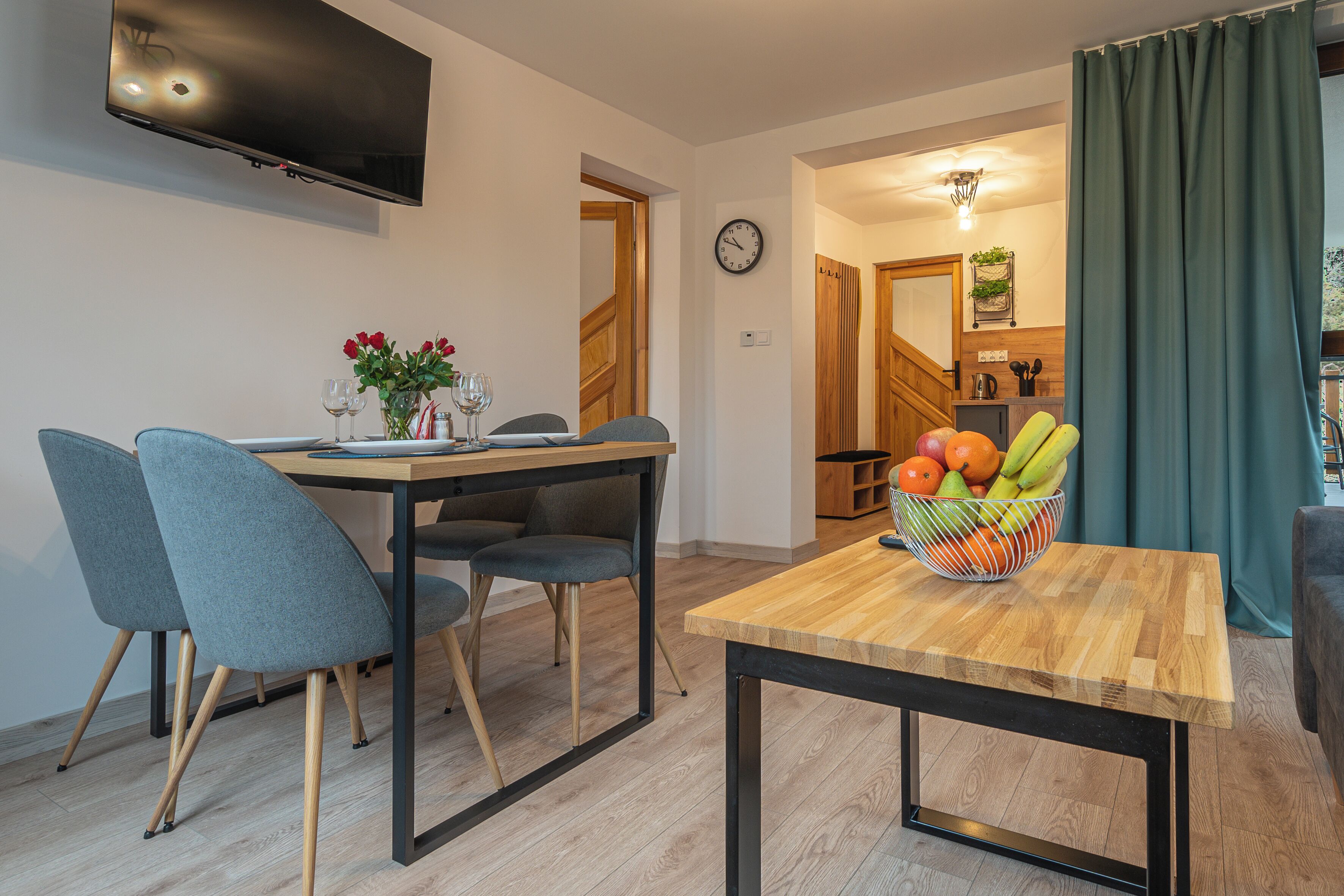 Un comedor con mesa de madera, sillas grises, un frutero y un televisor montado en la pared.