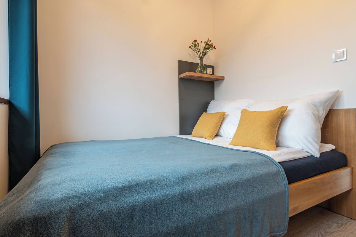 Ein ordentlich bezogenes Bett mit blauer Bettdecke, zwei Kissen und einem kleinen Regal mit einer Blumenvase.