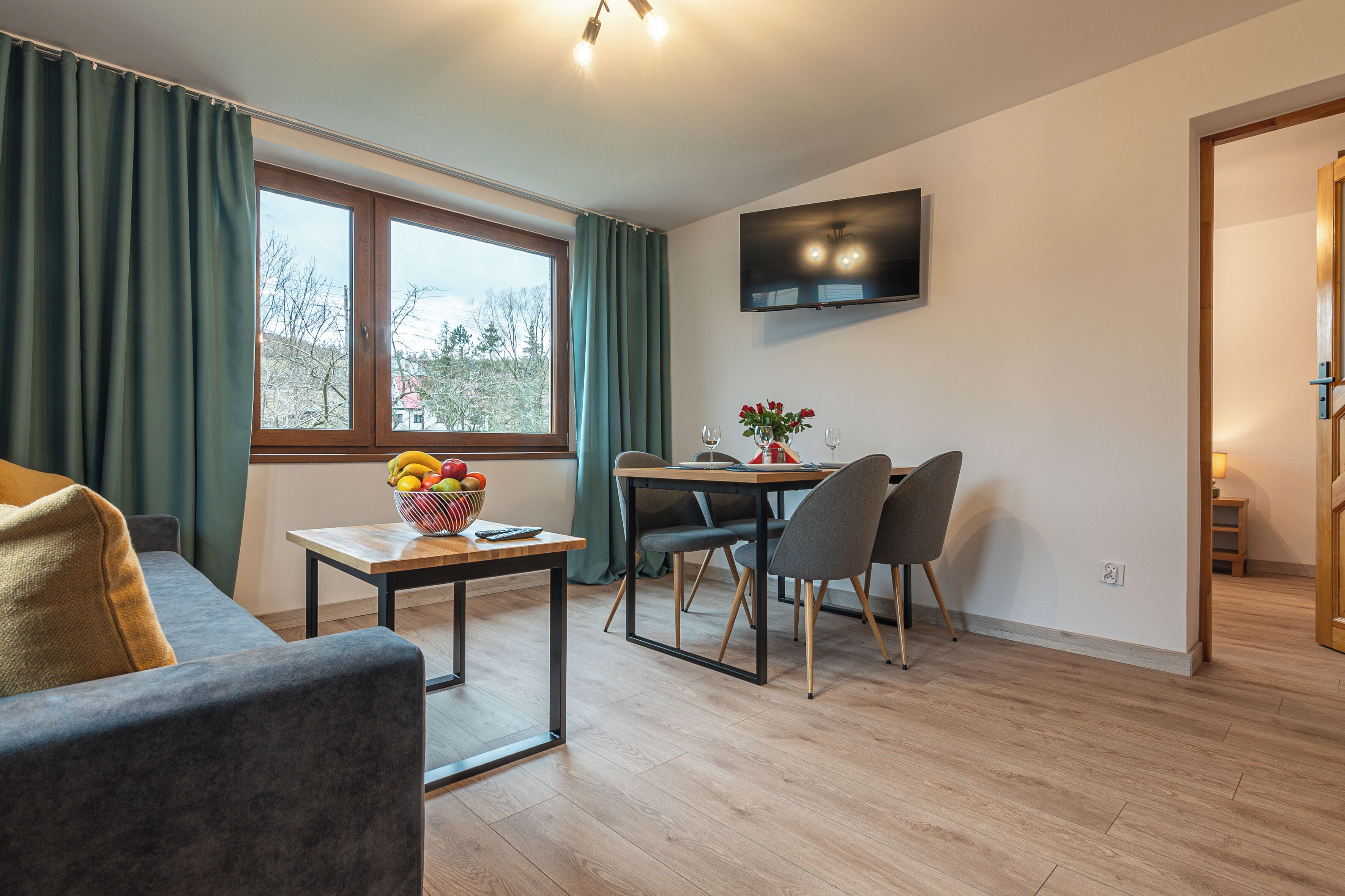 Ein modernes Wohnzimmer mit einer Couch, einem Esstisch mit Stühlen, einem an der Wand montierten Fernseher und einem Fenster mit Vorhängen.