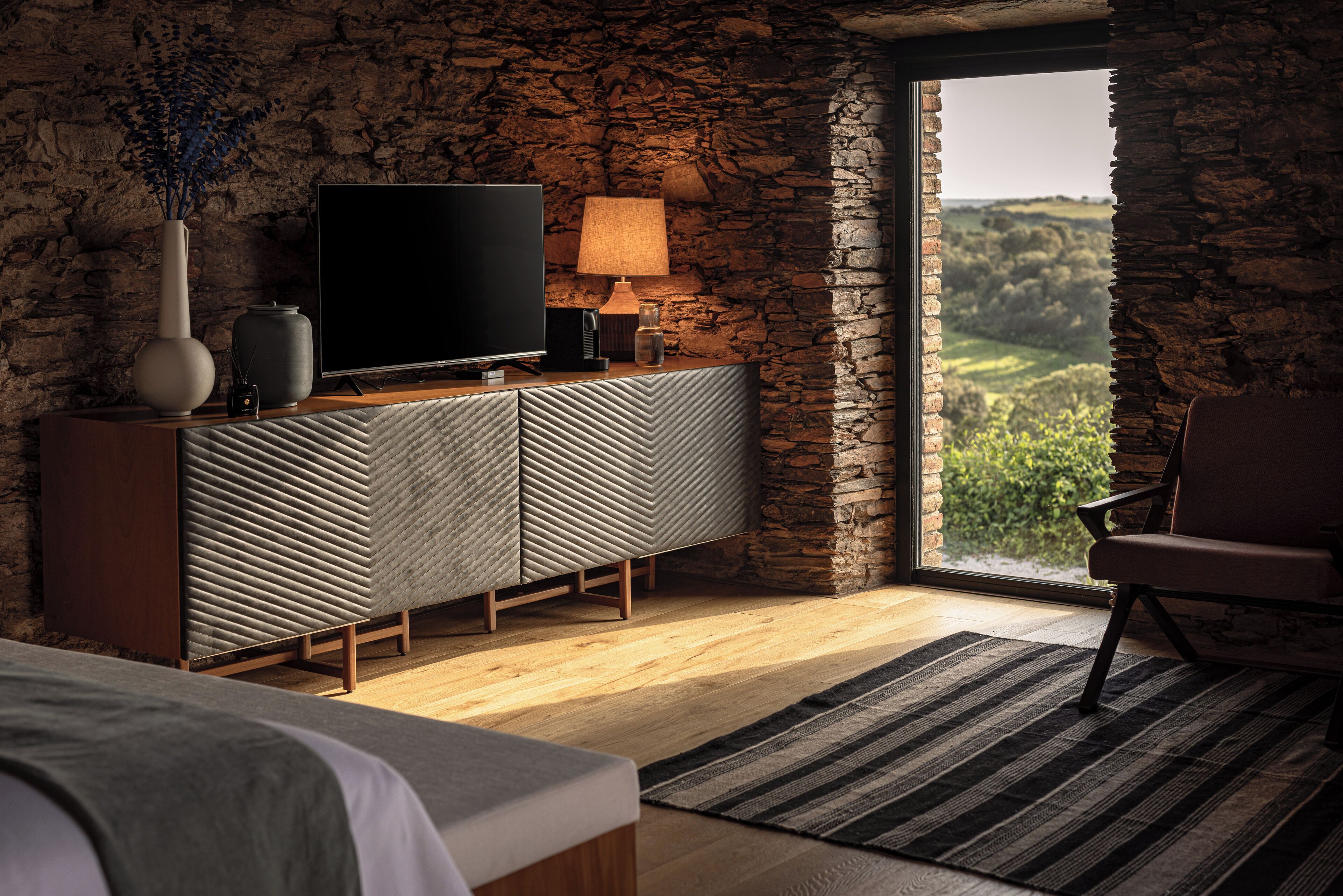 A modern bedroom with a stone wall, a television, a lamp, and a view of a green landscape.