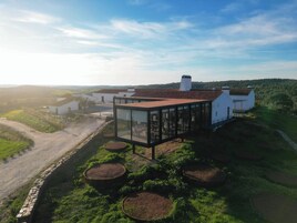 Una casa moderna con un invernadero de vidrio, rodeada por un muro de piedra y macetas circulares.