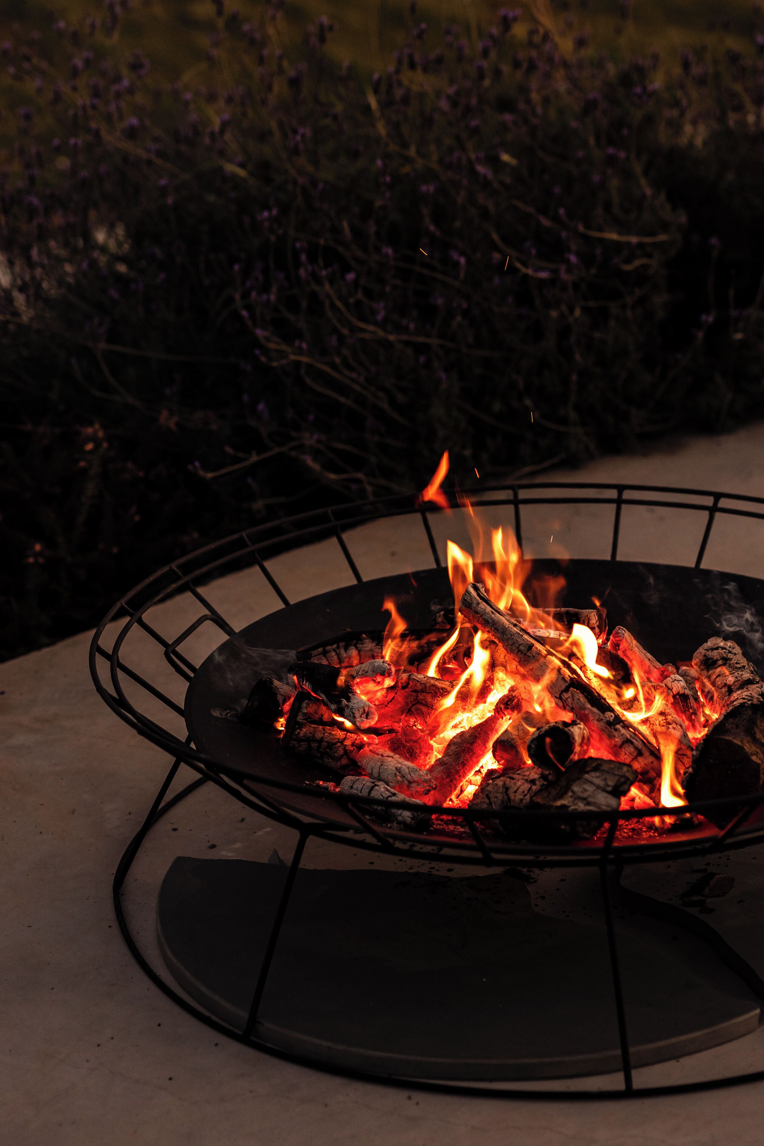 A circular outdoor fire pit with burning logs and flames, surrounded by a metal grate.
