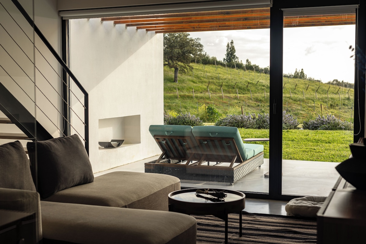 Un salón moderno con sofá, una mesa de centro y vistas a un paisaje verde a través de grandes puertas de vidrio.