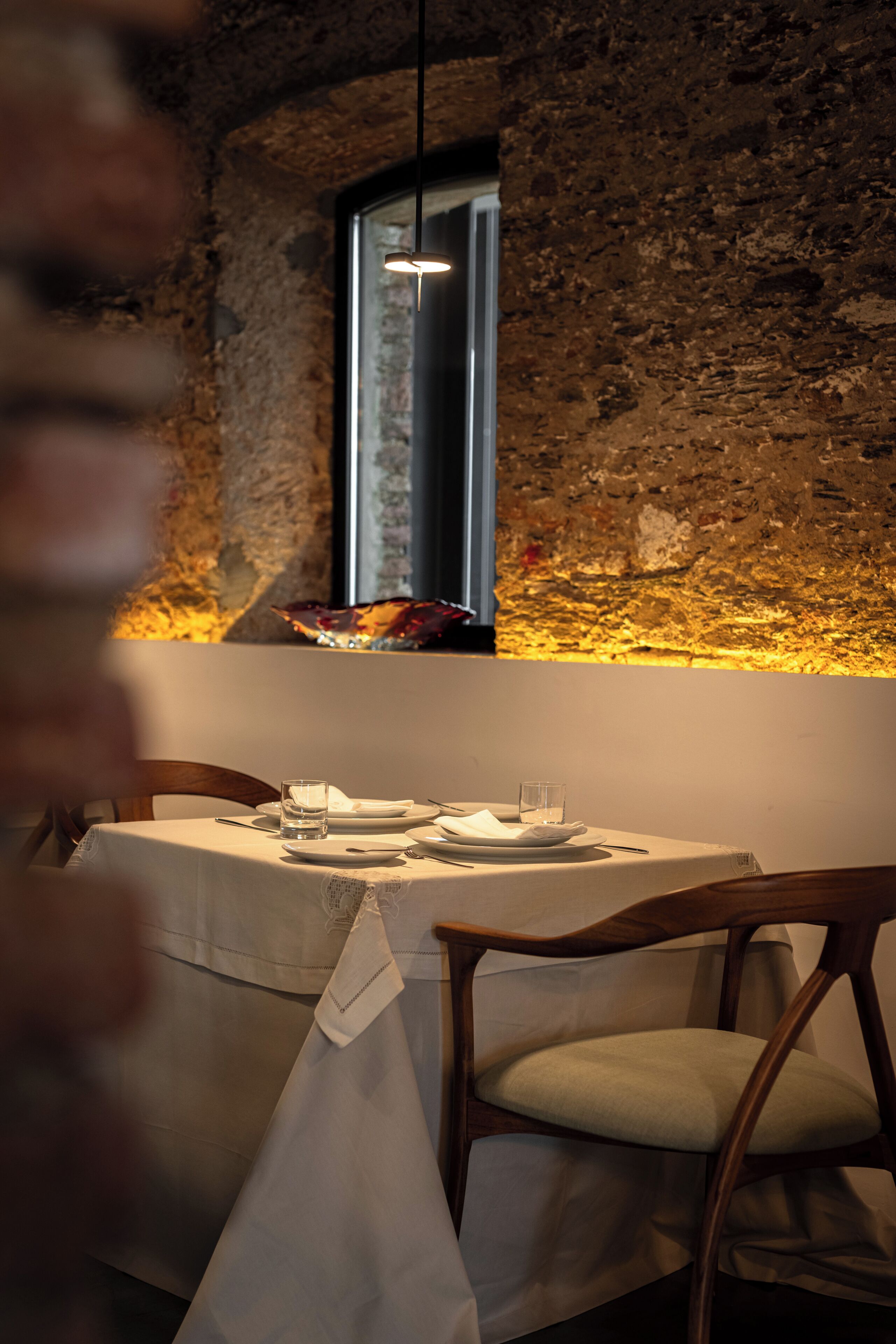 Une table à manger dressée avec des assiettes, des verres et une serviette, dans une pièce dotée d’un mur en pierre rustique et d’un luminaire suspendu.