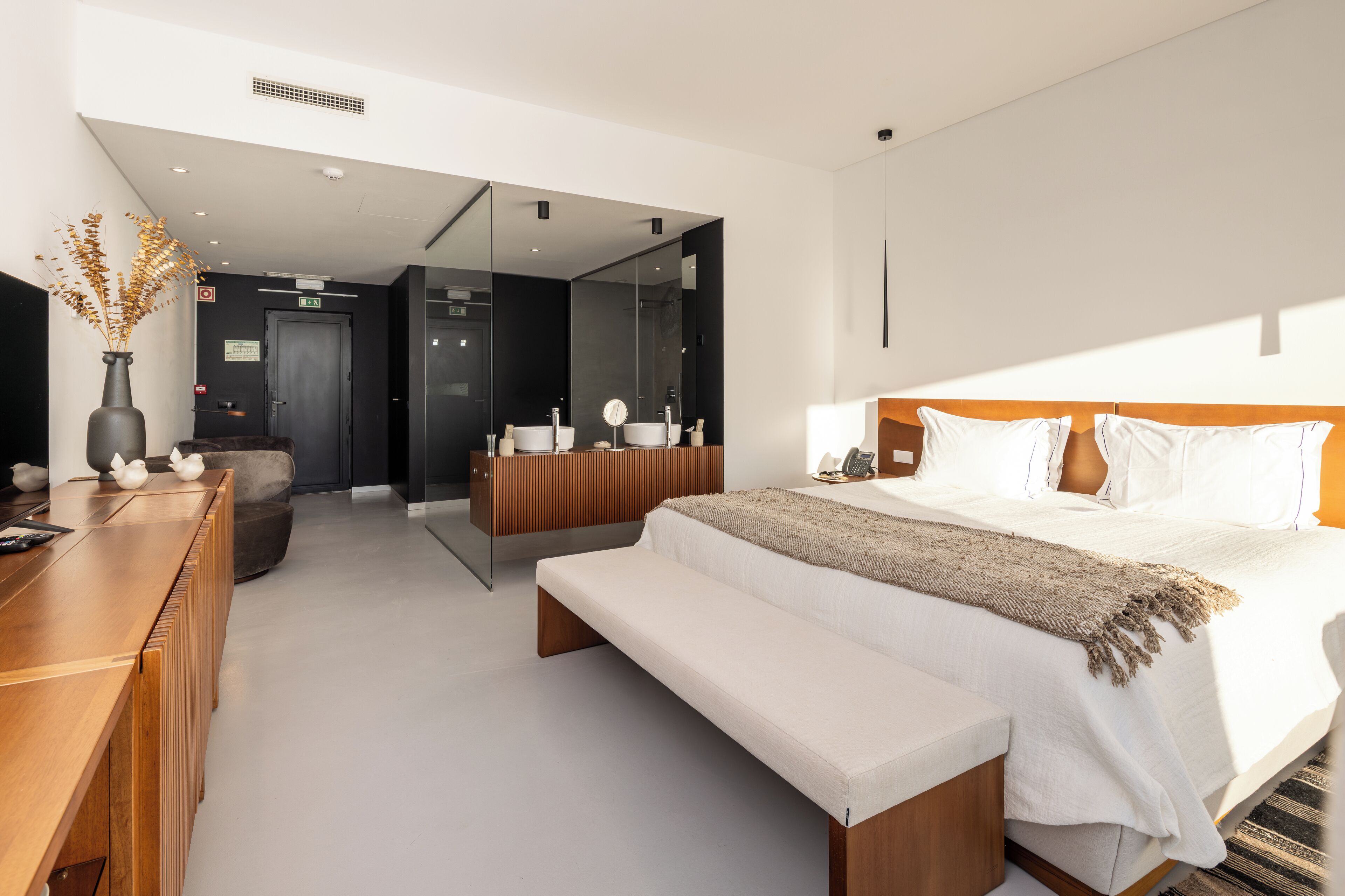 Een moderne slaapkamer met een groot bed, nachtkastje en een bankje. Er is een houten bureau en een vaas met gedroogde planten.