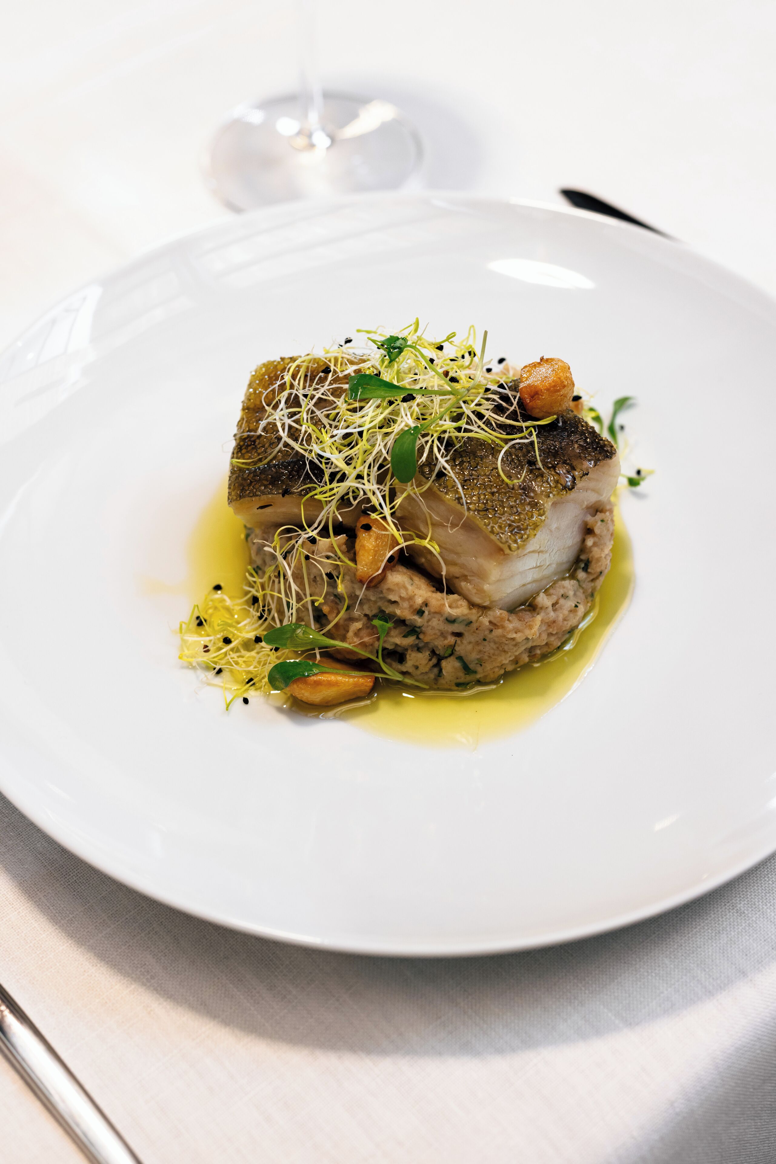 Une assiette blanche contenant un plat de poisson, agrémenté de verdure et de jeunes pousses, posée sur une nappe blanche.