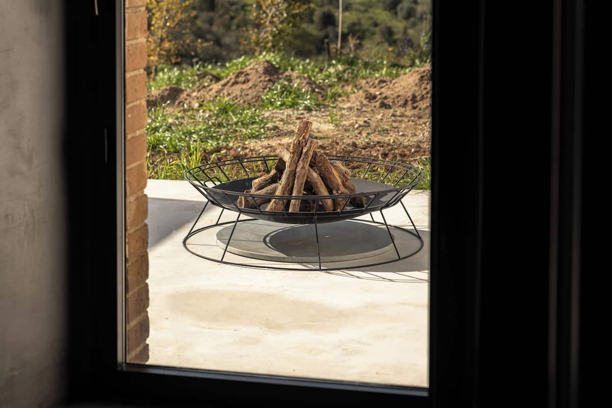 Uitzicht door een raam op een vuurkorf met hout, een betonnen terras en een glooiend landschap met bomen.