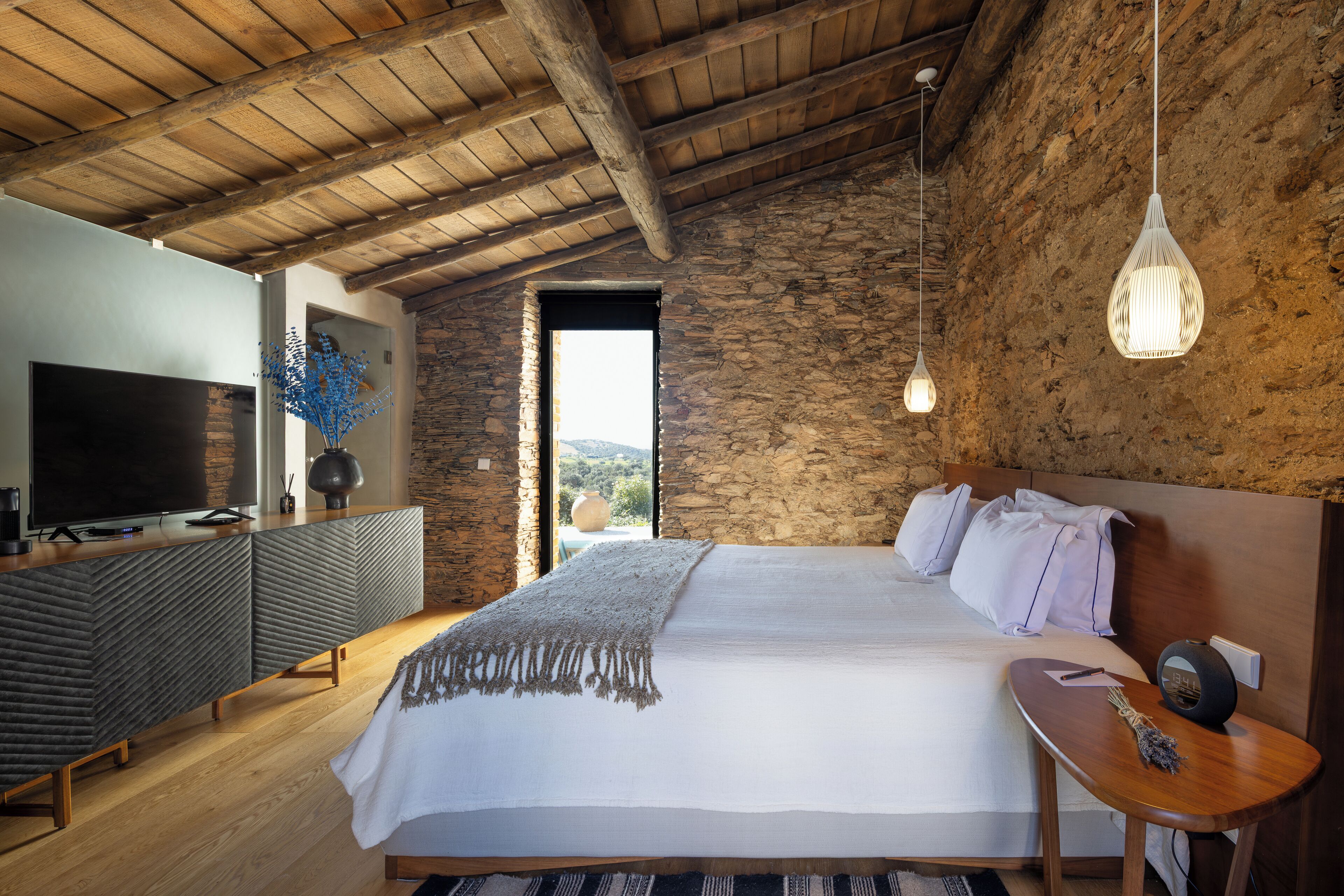 A bedroom with a large bed, a TV, a stone wall, and a view of the outdoors.