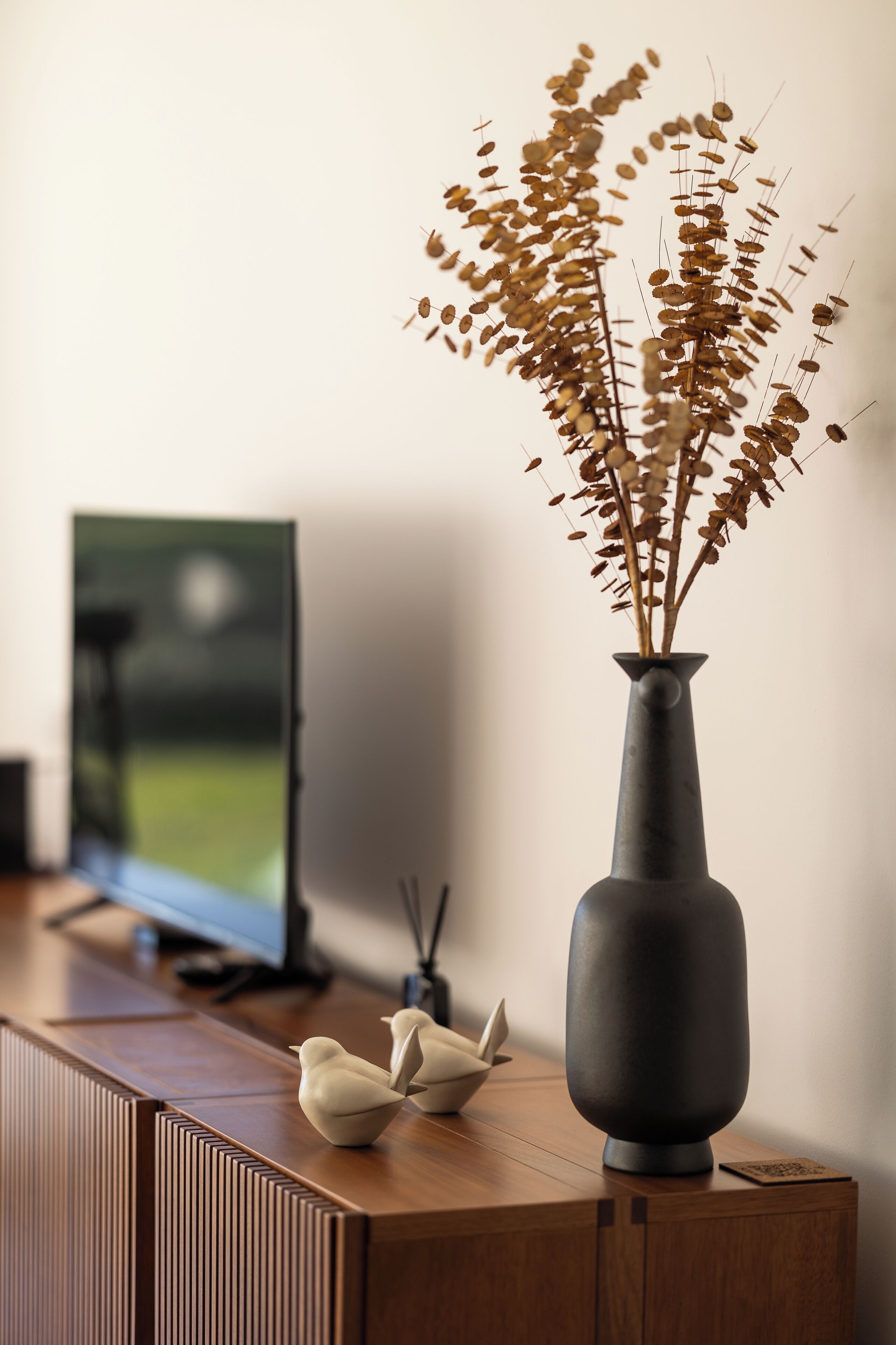 A wooden console table with a vase of dried flowers, a television, and decorative bird figurines.