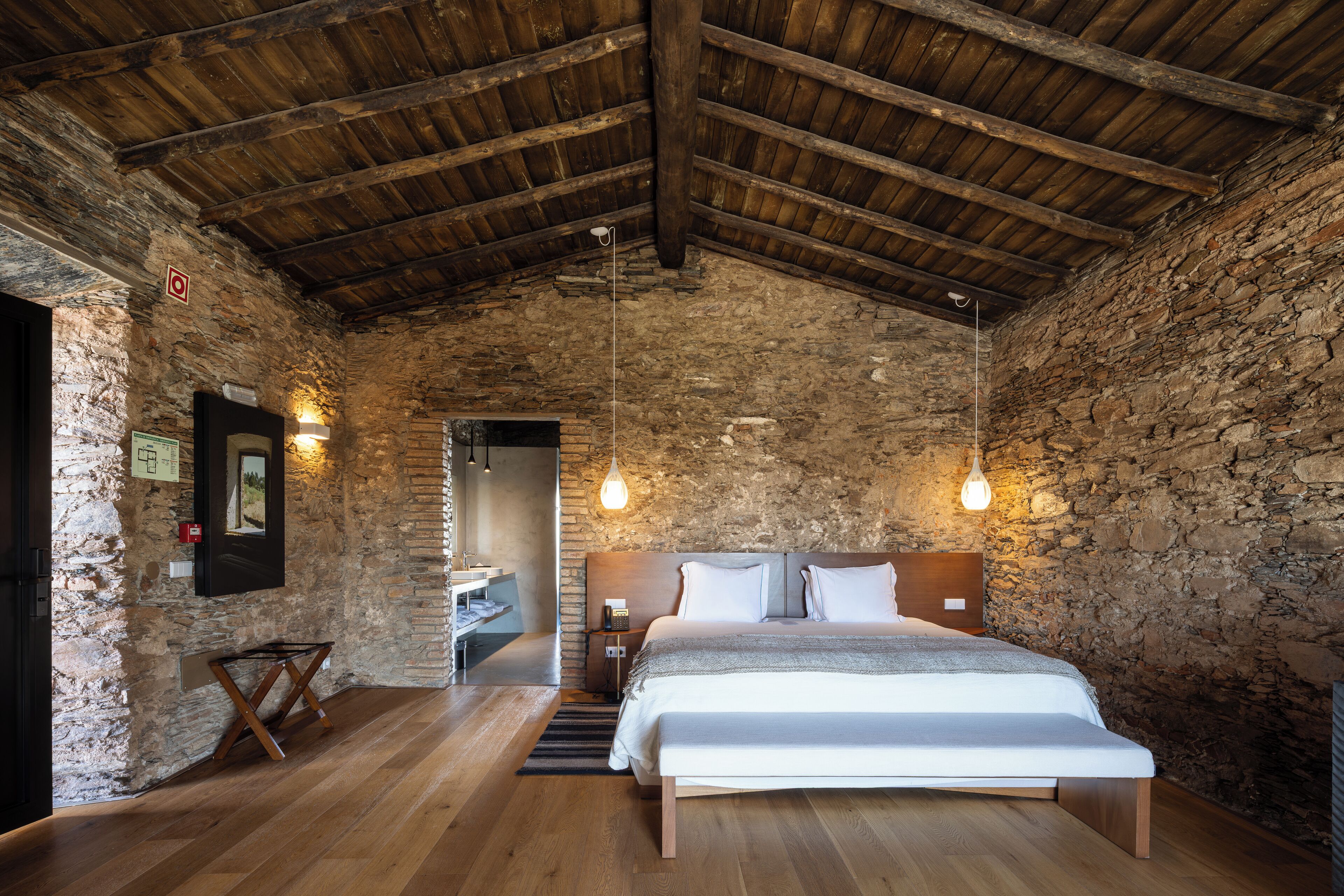 Ein rustikales Schlafzimmer mit einer Steinwand, Holzbalkendecke, einem Bett mit weißer Bettwäsche und hängenden Pendelleuchten.